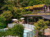 Spa-Bereich mit Pool, Sonnenschirmen und Sitzbereichen, umgeben von viel Grün; im Hintergrund rustikale Holzhäuser mit begrünten Dächern.