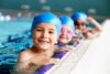 Kinder mit Badekappen am Beckenrand, lachend in die Kamera schauend.