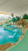 Innenaufnahme eines großen Erlebnisbeckens mit türkisfarbenem Wasser, Felsenlandschaft und grünen Wasserrutschen, die von einer Galerie aus starten.