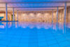 Innenaufnahme eines weitläufigen Schwimmbeckens mit blauer Beleuchtung, weißen Säulen und heller Decke.