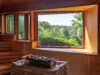 Sauna mit großem Panoramafenster, das den Blick auf grüne Bäume und Landschaft freigibt, im Vordergrund ein Saunaofen mit Steinen.