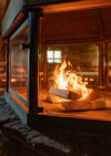 Sauna mit zentralem Kamin, in dem offenes Feuer hinter Glas lodert, umgeben von Holzbänken.