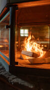 Sauna mit zentralem Kamin, in dem offenes Feuer hinter Glas lodert, umgeben von Holzbänken.